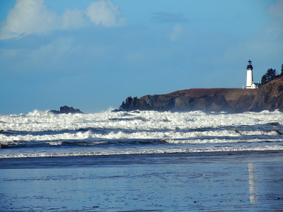 [The lighthouse seems to sit back from the edge of the rock which is being hit by powerful, frothy waves. This view was taken from a nearby beach.]